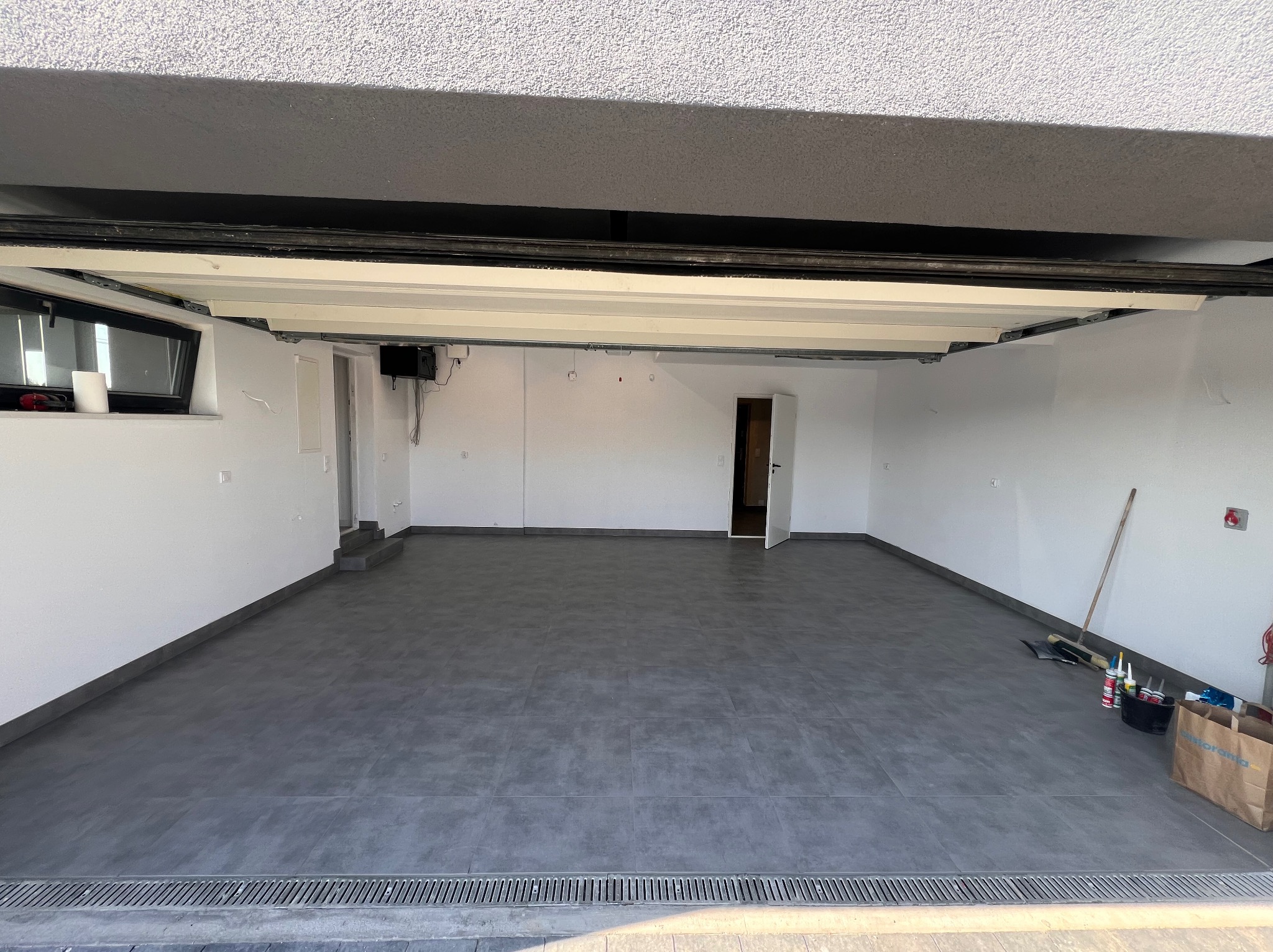 Wykończony garaż z szarą podłogą z płytek, białe ściany, widoczne narzędzia i materiały budowlane w rogu, odpływ liniowy przy wjeździe.