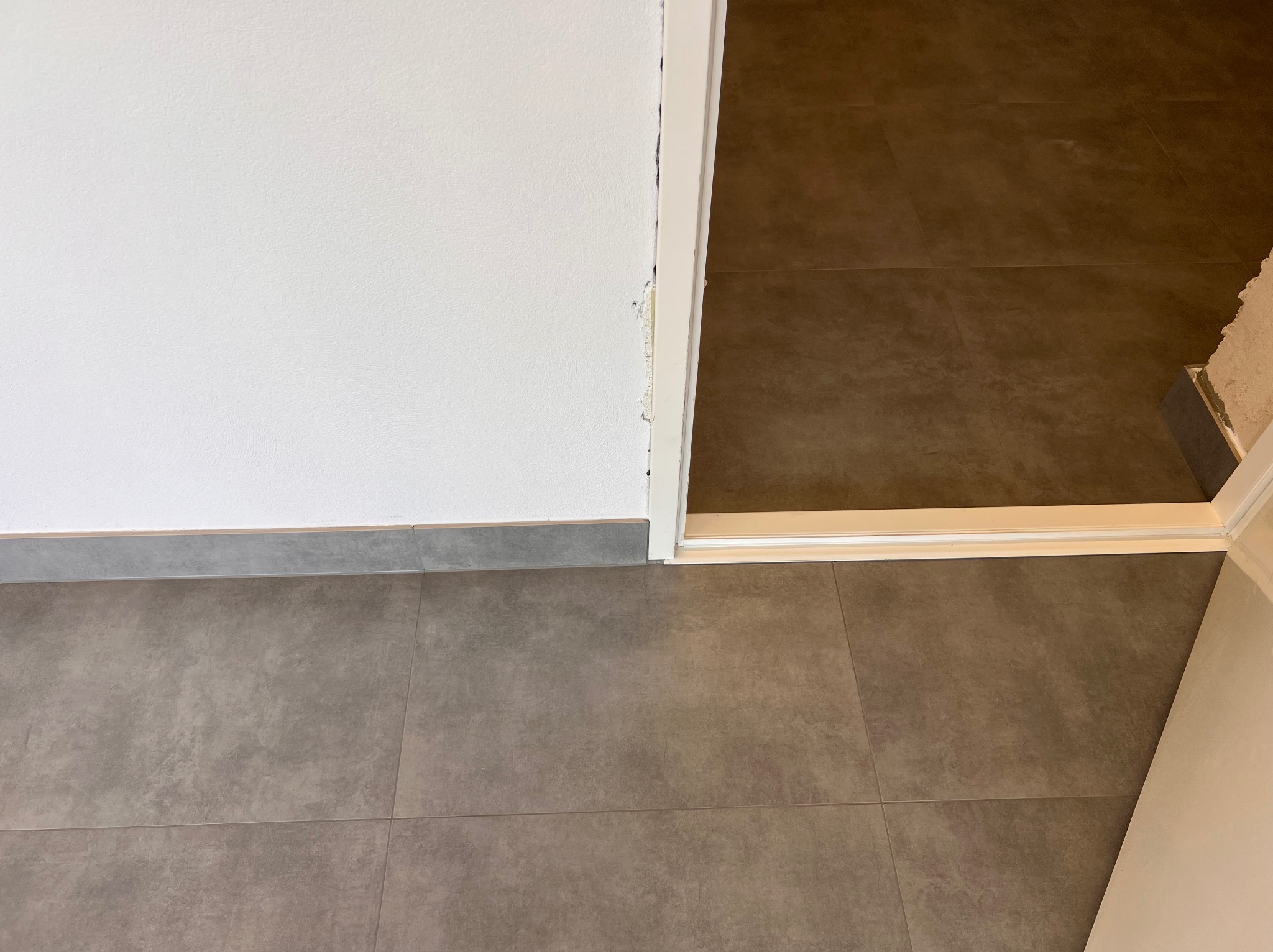Szare płytki podłogowe w nowym pomieszczeniu, widoczny fragment ściany i próg drzwiowy z niedokończonym wykończeniem.