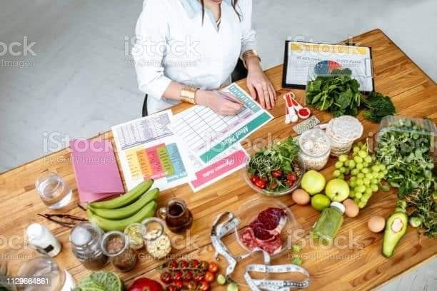Kobieta w białym uniformie wypełnia plan posiłków na drewnianym stole, otoczona świeżymi produktami: warzywami, owocami, mięsem, ziarnami i przyprawami w słoiczkach.