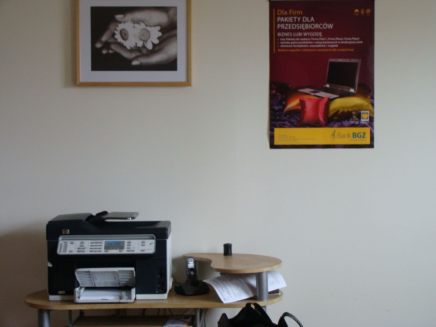 Plakat reklamowy Banku BGZ z ofertą dla firm, wiszący obok czarno-białej fotografii rąk trzymających kwiaty, poniżej stoi drukarka wielofunkcyjna i telefon na drewnianym stoliku.