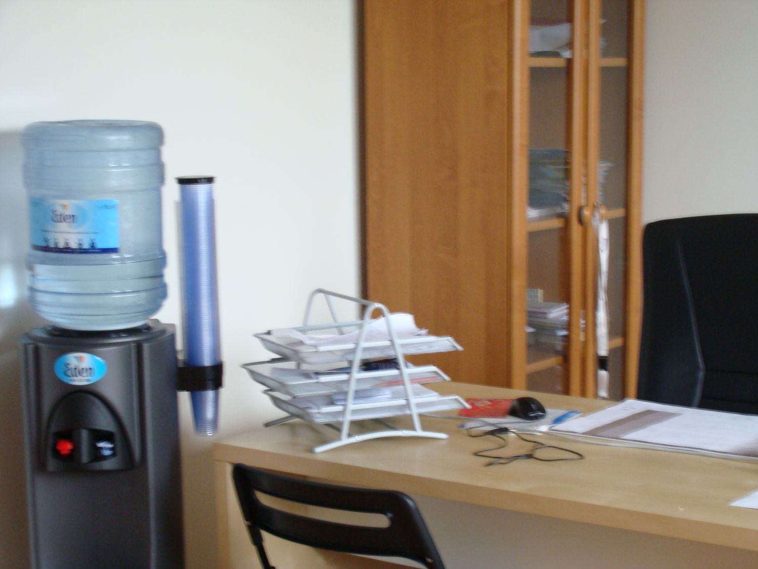 Biuro: dystrybutor wody, biurko z dokumentami i organizerem na papiery, w tle szafka z przeszklonymi drzwiami i czarne krzesło biurowe.