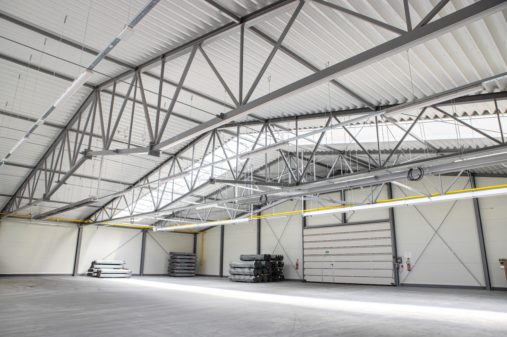 Wnętrze nowej hali z widoczną konstrukcją stalową dachu, ścianami z paneli i bramą segmentową, przygotowane do użytku, ze stertami materiałów budowlanych na podłodze.