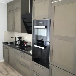 Nowoczesna kuchnia z szarymi drewnianymi szafkami, czarnym blatem i wbudowanym piekarnikiem Miele, z widocznym fragmentem podłogi z paneli imitujących drewno.