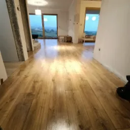 Nowo ułożona, błyszcząca podłoga z paneli imitujących drewno w przestronnym, jasnym wnętrzu, z widokiem na krajobraz za oknem.