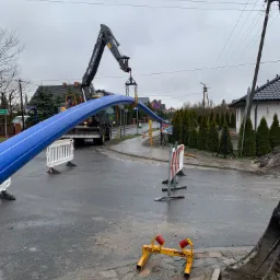 Ciężki dźwig Volvo podnosi niebieską rurę na mokrej ulicy w pochmurny dzień, widoczne znaki ostrzegawcze i zabudowa jednorodzinna w tle.