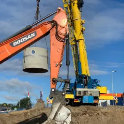 Pomarańczowy dźwig Doosan podnosi betonowy krąg z otworem, na pierwszym planie łyżka koparki, w tle żółto-niebieski dźwig, widoczne rury w wykopie.