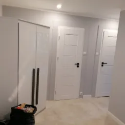 Szare ściany świeżo po malowaniu, widoczne białe drzwi z czarnymi klamkami i gładka podłoga, przy ścianie stoi biała szafa z ciemnymi uchwytami, a obok niej czarny worek ze śmieciami po remoncie.