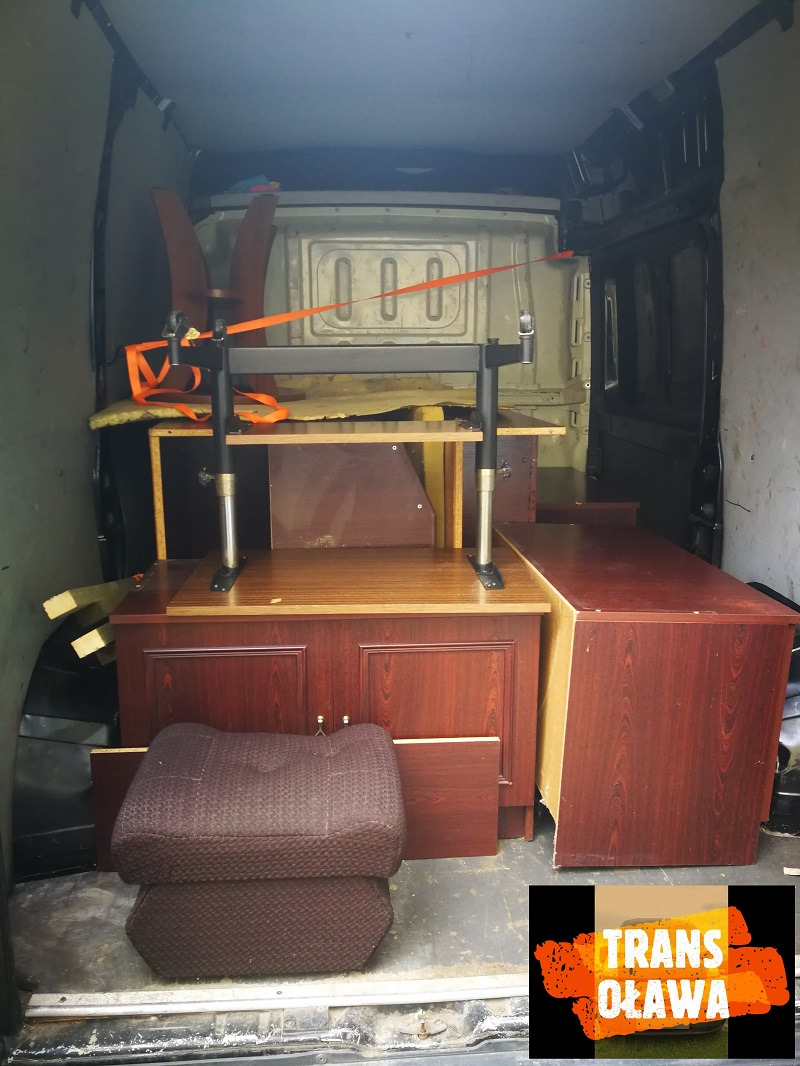 Wnętrze busa załadowane meblami w kolorze ciemnego mahoniu, zabezpieczonymi pasami transportowymi, gotowymi do przewozu, z widocznym logiem firmy transportowej w prawym dolnym rogu.