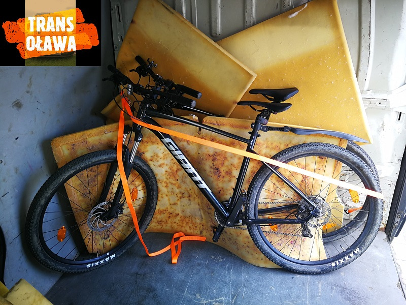 Rower Giant zabezpieczony pomarańczowym pasem transportowym wewnątrz furgonetki, otoczony dużymi arkuszami pianki o nieregularnych kształtach i żółtej barwie, gotowy do transportu.