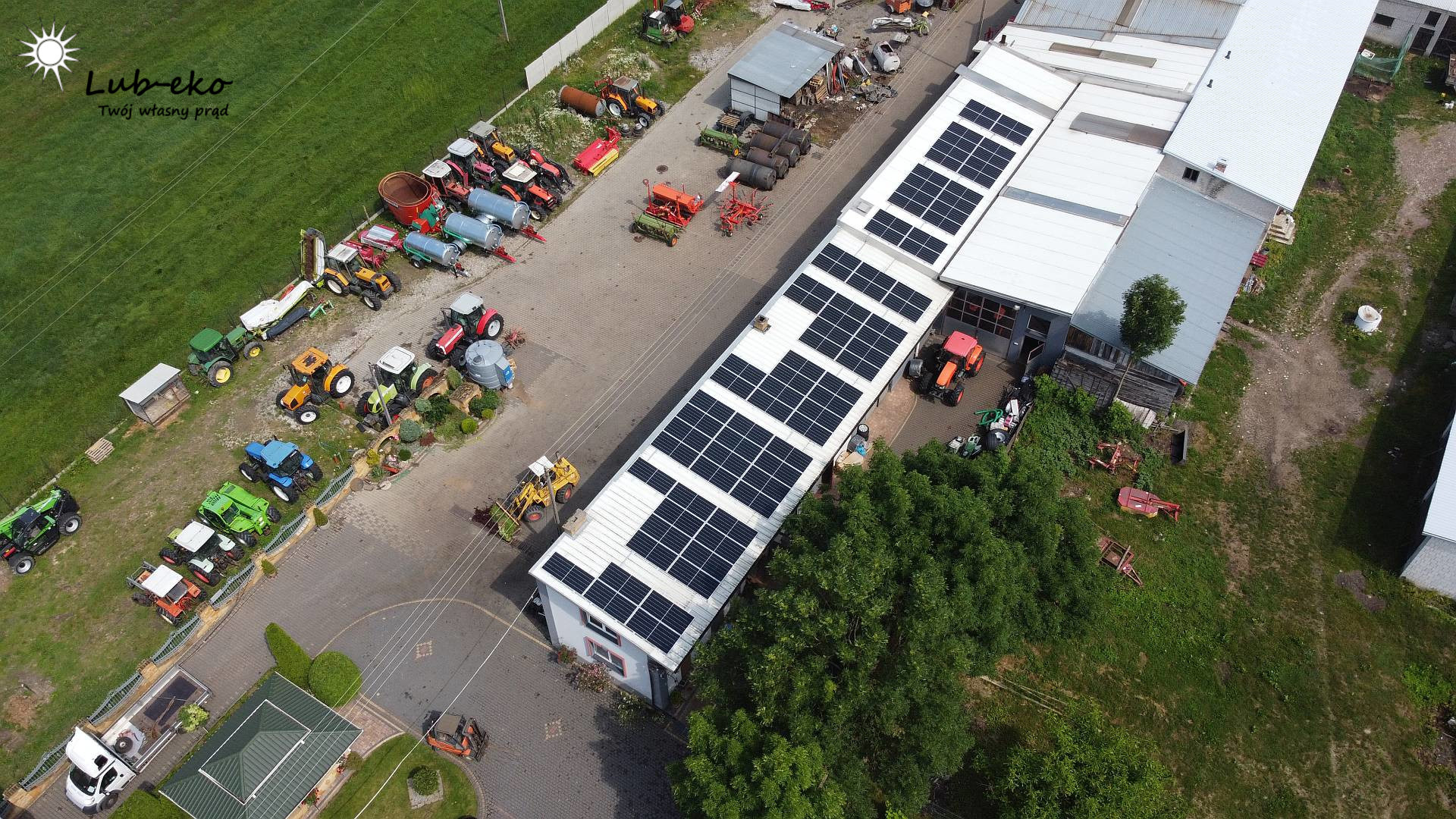 Widok z góry na budynek z zainstalowanymi panelami fotowoltaicznymi na dachu, otoczony zielenią i maszynami rolniczymi, z logo firmy w lewym górnym rogu.