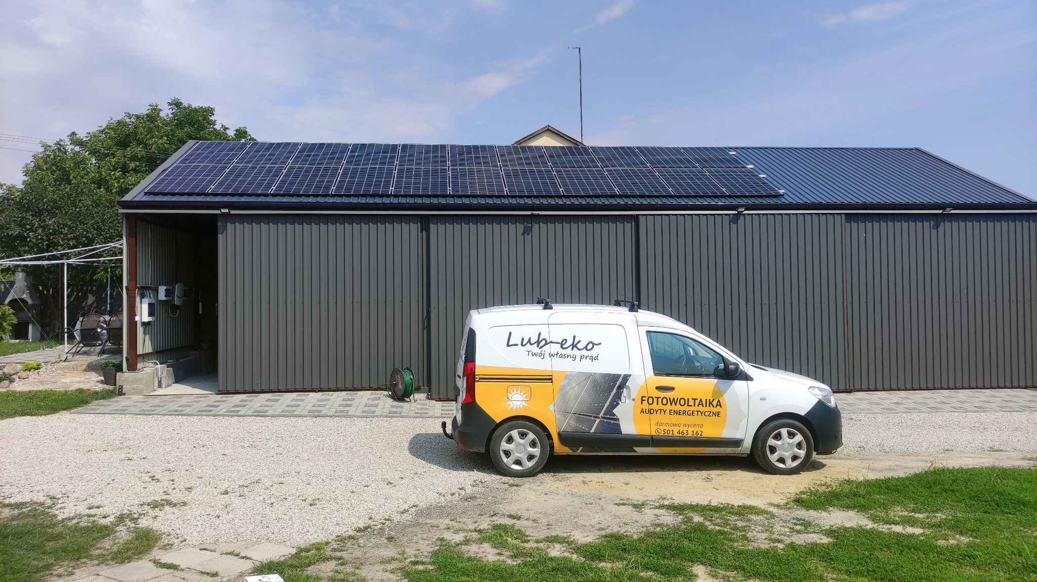 Instalacja fotowoltaiczna na dachu garażu w Lublinie, z zaparkowanym obok samochodem firmowym z logo i informacjami kontaktowymi.