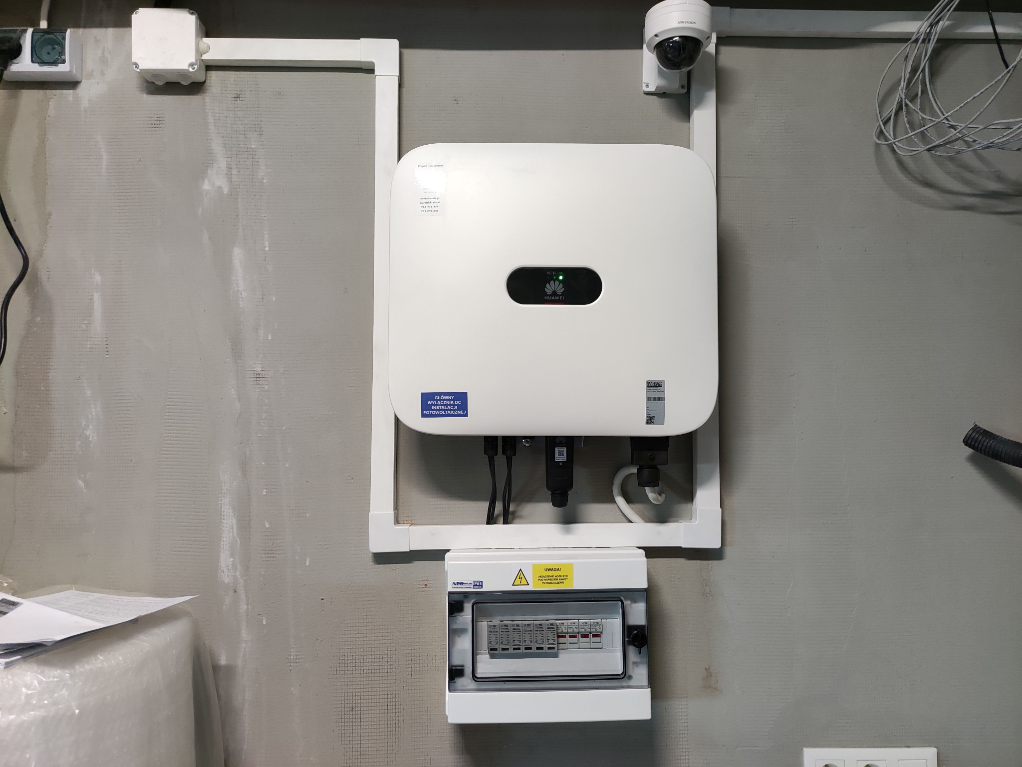 Biały falownik Huawei z podłączonymi kablami i wyłącznikiem DC instalacji fotowoltaicznej, zamontowany na szarej ścianie z widoczną kamerą Hikvision pod sufitem.