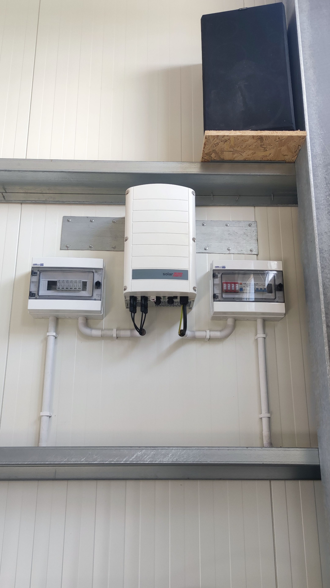 Biały falownik SolarEdge z podłączonymi kablami i dwiema skrzynkami bezpiecznikowymi zamontowany na białej ścianie z paneli, widoczne szyny montażowe i czarna skrzynka na górnej półce.