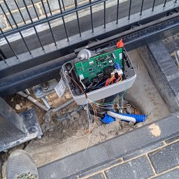 MIK-ELEKTRO Kamil Lipka - Otwarta obudowa automatu bramy przesuwnej z widoczną płytą sterującą, przewodami i elementami montażowymi, umieszczona w wykopie obok metalowej bramy, z perspektywy osoby stojącej nad nią w szarych...