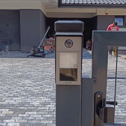 MIK-ELEKTRO Kamil Lipka - Szary wideodomofon Hikvision zamontowany na słupku furtki, widoczna kostka brukowa na podjeździe i pracownik układający ją w tle.