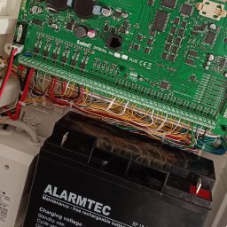 MIK-ELEKTRO Kamil Lipka - Wnętrze skrzynki alarmowej z widoczną płytą główną Satel Integra 64 Plus, podłączonymi przewodami oraz akumulatorem Alarmtec BP 18-12. Na dolnej krawędzi widoczne opakowania po wkrętach Cobalt...