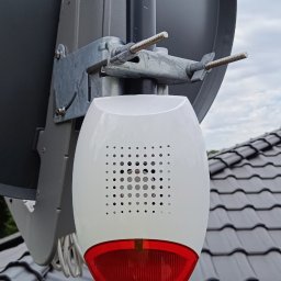 MIK-ELEKTRO Kamil Lipka - Zewnętrzna, biało-czerwona syrena alarmowa z perforowaną obudową, zamontowana na metalowym słupku obok anteny satelitarnej na tle dachu z szarej dachówki.