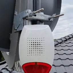 Zewnętrzna, biało-czerwona syrena alarmowa z perforowaną obudową, zamontowana na metalowym słupku obok anteny satelitarnej na tle dachu z szarej dachówki.