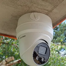 Kamera monitoringu marki Hikvision zamontowana pod sufitem na zewnątrz budynku, widok z dołu na tle zieleni.