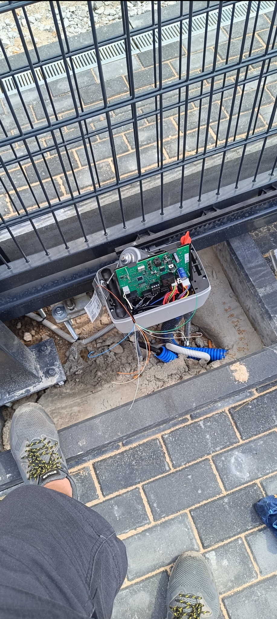 Otwarta obudowa automatu bramy przesuwnej z widoczną płytą sterującą, przewodami i elementami montażowymi, umieszczona w wykopie obok metalowej bramy, z perspektywy osoby stojącej nad nią w szarych...