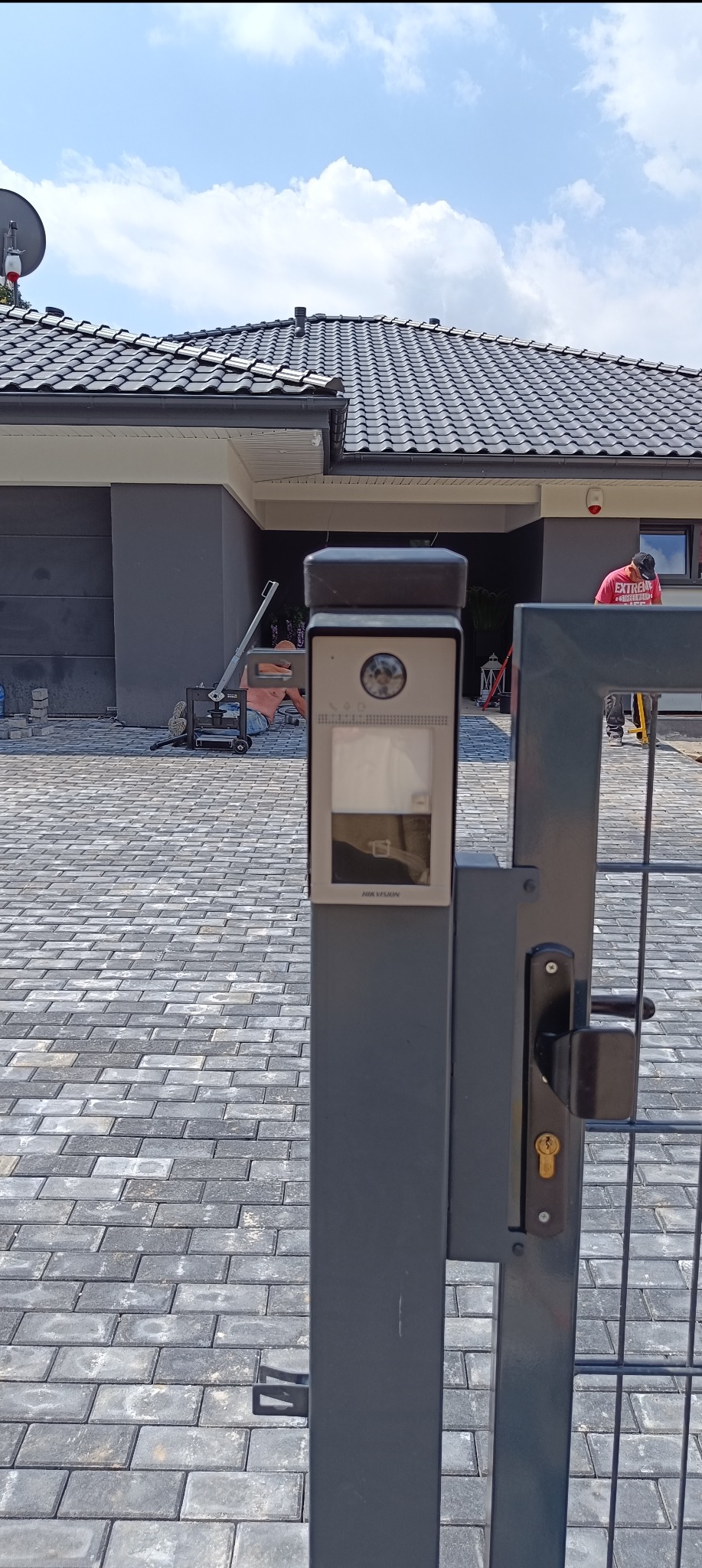 Szary wideodomofon Hikvision zamontowany na słupku furtki, widoczna kostka brukowa na podjeździe i pracownik układający ją w tle.