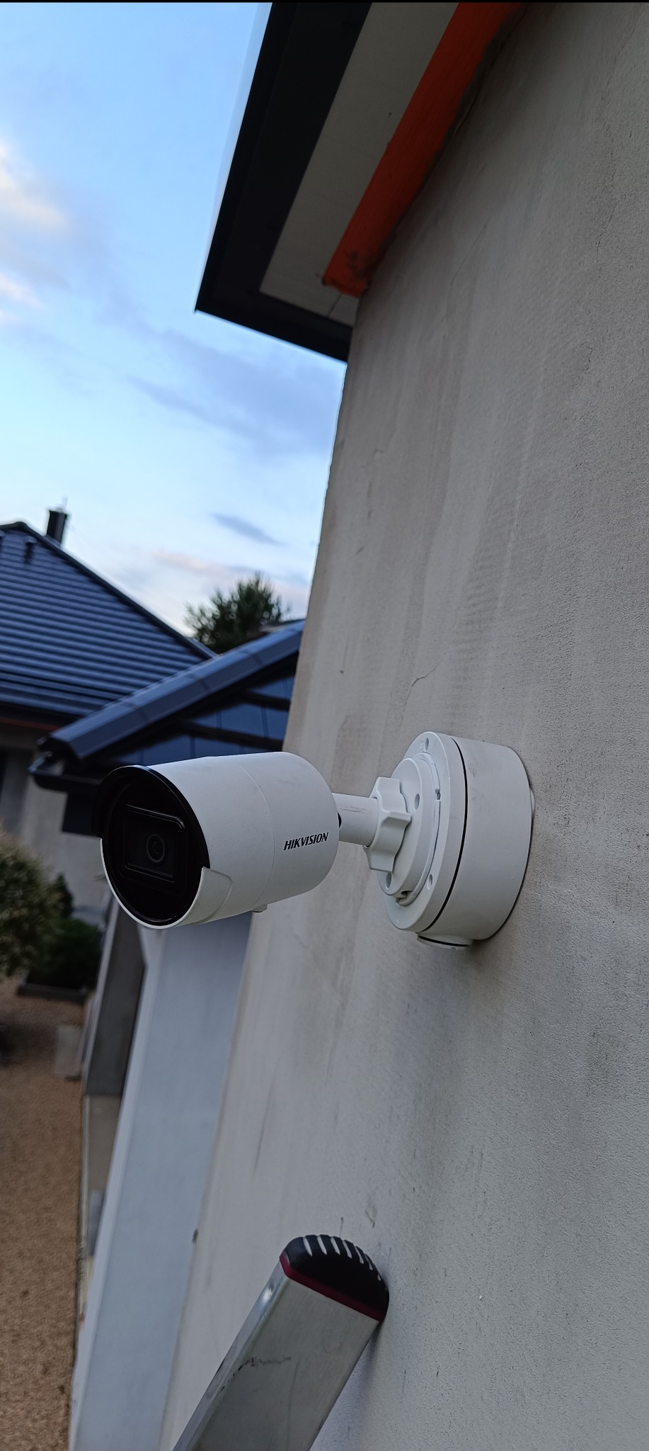 Biała kamera monitoringu Hikvision zamontowana na szarej ścianie budynku, widoczna drabina oparta o ścianę, w tle niebieskie niebo i fragment dachu