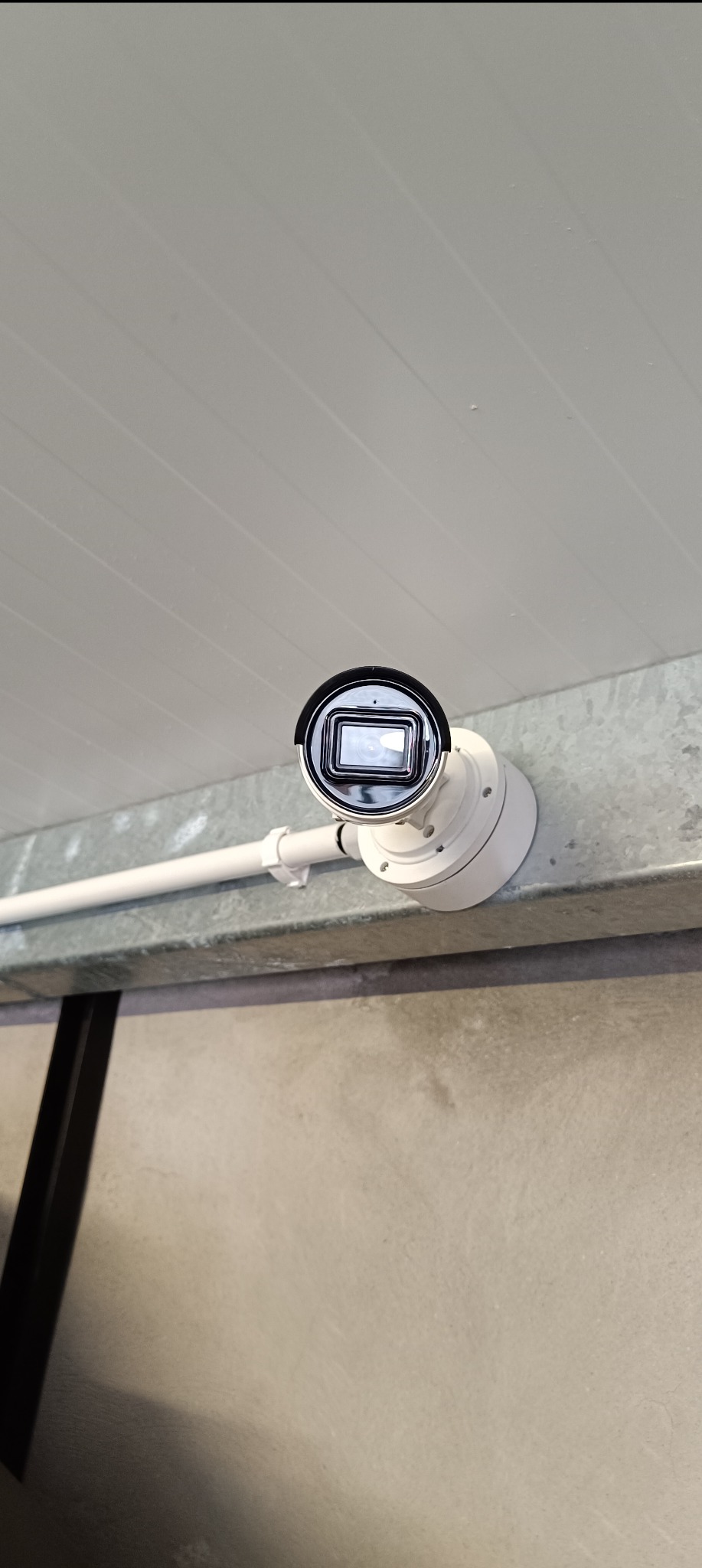 Biała kamera monitoringu zamontowana na metalowej szynie pod sufitem z widocznymi rurami instalacyjnymi.
