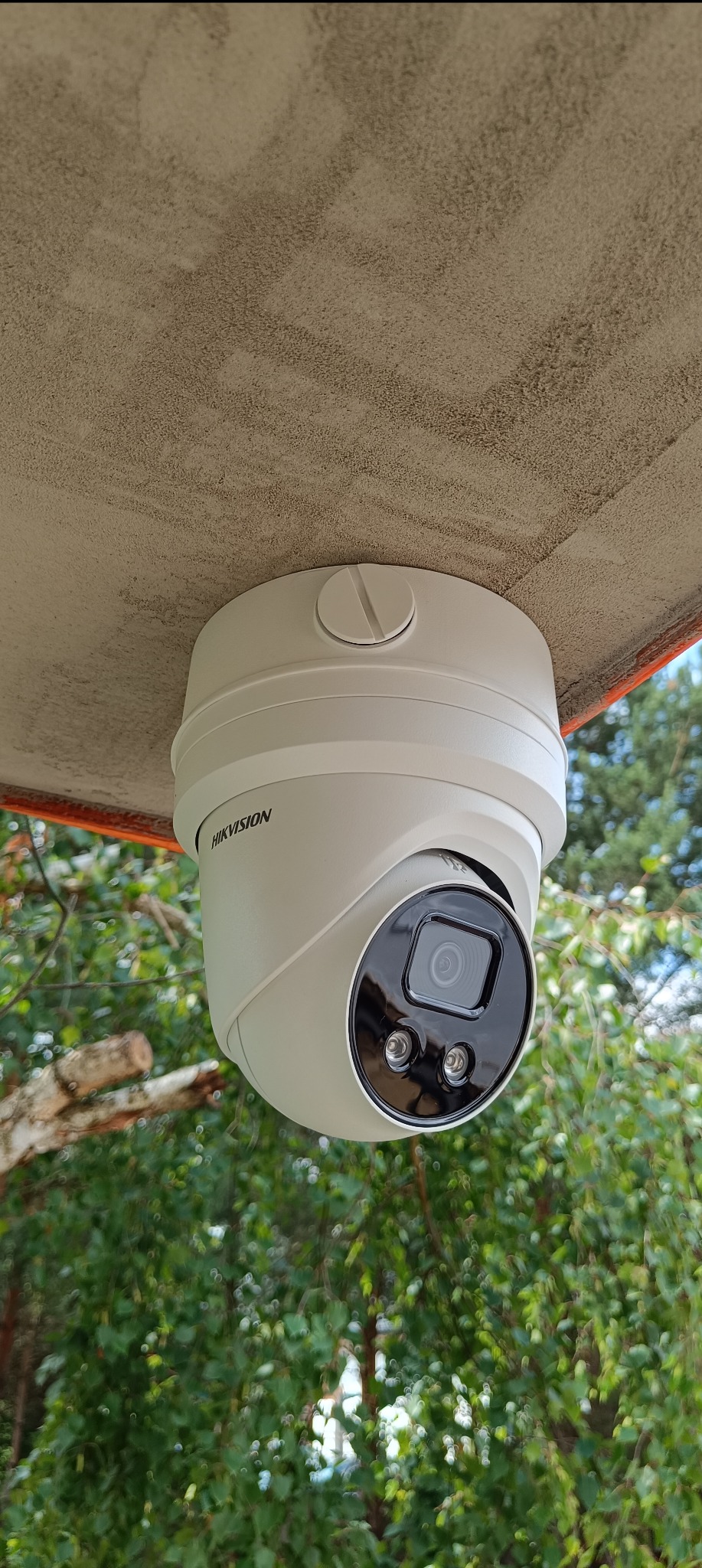 Kamera monitoringu marki Hikvision zamontowana pod sufitem na zewnątrz budynku, widok z dołu na tle zieleni.