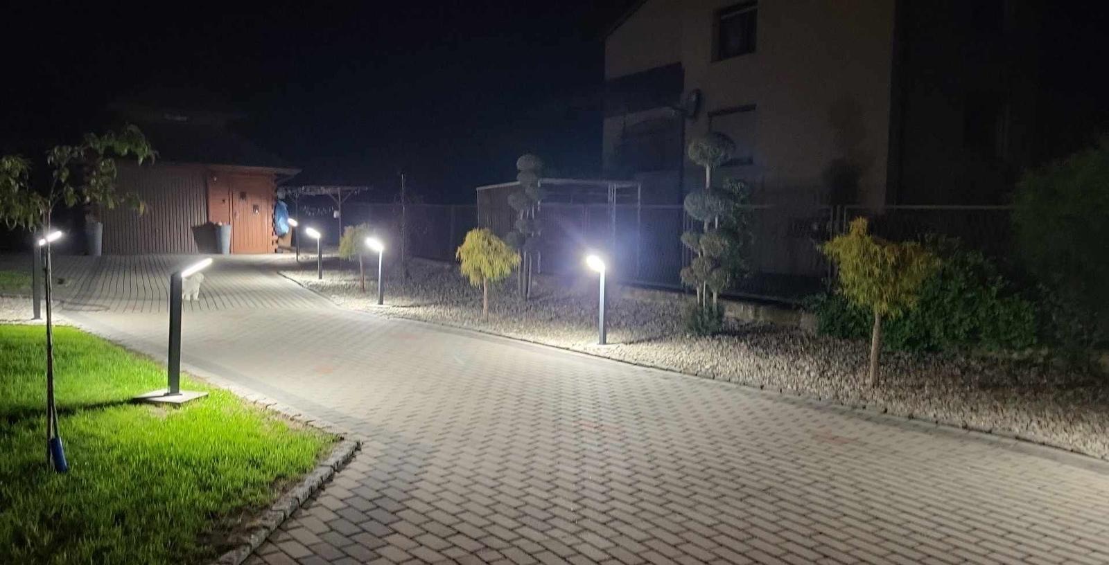 Oświetlona ścieżka z kostki brukowej prowadząca do domu nocą, z nowoczesnymi lampami ogrodowymi po bokach, podkreślającymi architekturę krajobrazu i roślinność.