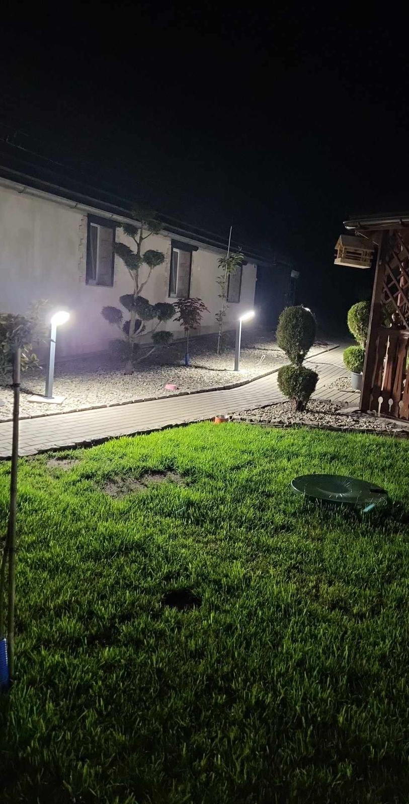Oświetlony ogród nocą z lampami stojącymi wzdłuż ścieżki, ozdobnymi krzewami i trawnikiem. Widoczne elementy architektury budynku w tle.