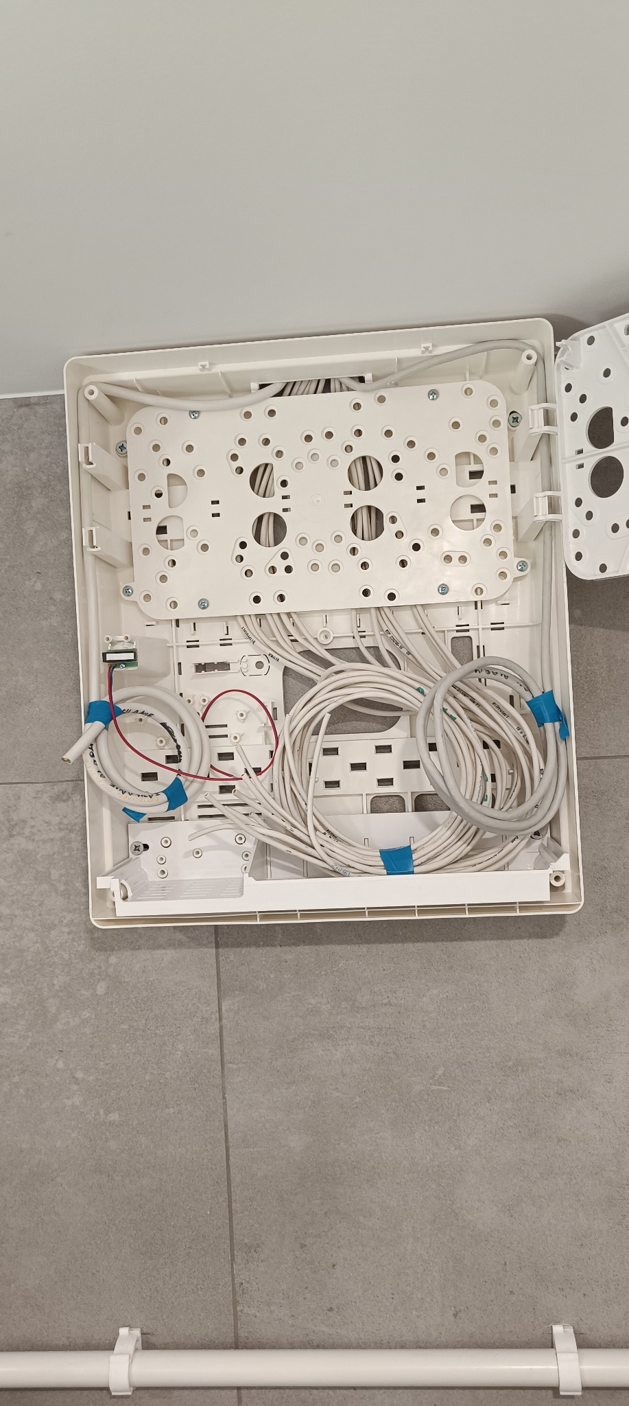 Otwarta, biała skrzynka rozdzielcza z uporządkowanymi przewodami elektrycznymi, zabezpieczonymi niebieską taśmą, widok z góry na szare płytki podłogowe.
