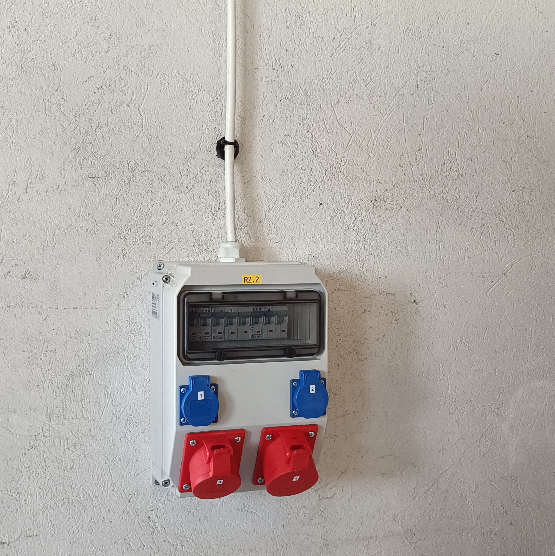 Rozdzielnica elektryczna z zabezpieczeniami i gniazdami siłowymi (czerwone i niebieskie) zamontowana na szarej, chropowatej ścianie. Widoczna rurka instalacyjna przymocowana uchwytem.