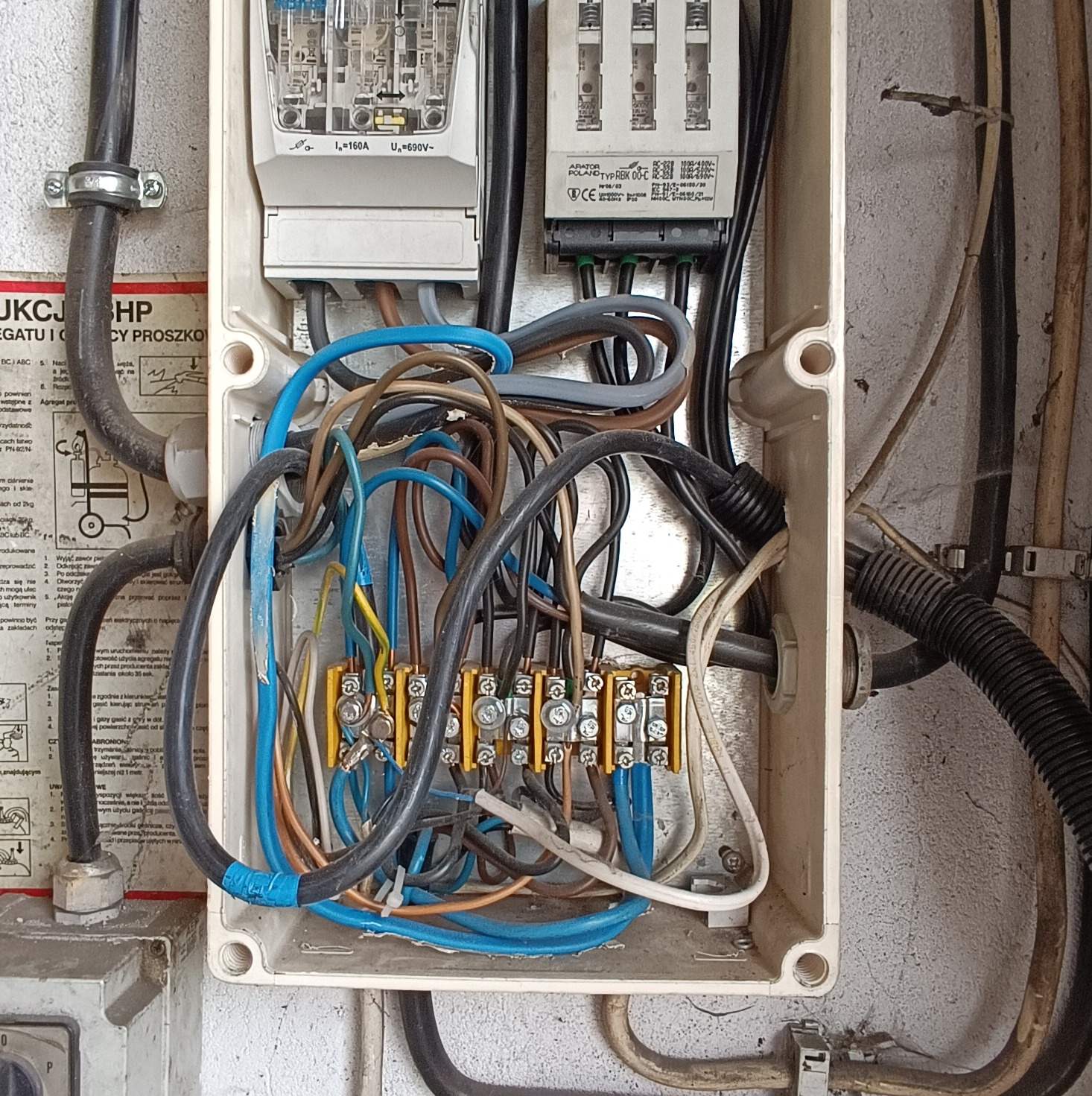 Otwarta skrzynka elektryczna z widocznymi przewodami, bezpiecznikami i listwami zaciskowymi, niektóre przewody owinięte niebieską taśmą izolacyjną.