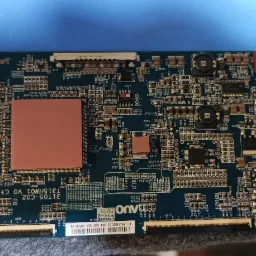 Zdjęcie niebieskiej płyty głównej telewizora z widocznymi elementami elektronicznymi, w tym układami scalonymi, kondensatorami i rezystorami, z bliska na niebieskim podłożu.