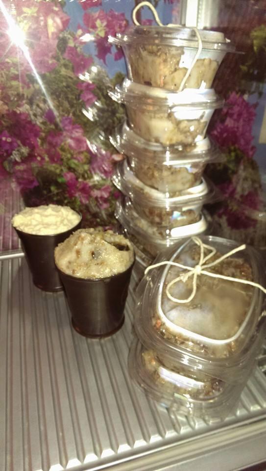 Desery w plastikowych pojemnikach i brązowych kubeczkach na metalowej półce, z kwiatami w tle. Domowe wyroby cukiernicze gotowe do sprzedaży.