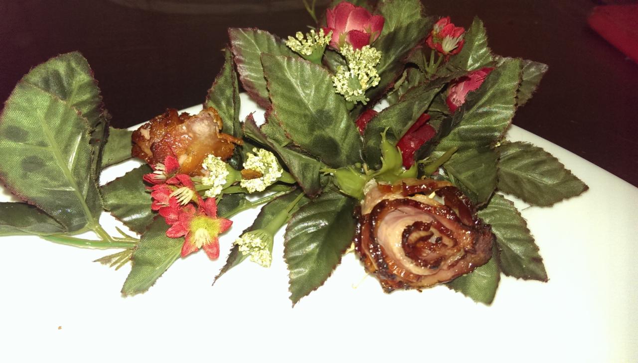 Dekoracja stołu: róża z boczku otoczona sztucznymi kwiatami i liśćmi na białym talerzu, ciemne tło. Nietypowa kompozycja kulinarna.