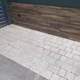 S/Mdd-Bud - Fragment elewacji budynku z ciemnozielonym tynkiem i panelami imitującymi drewno, przylegający do chodnika wyłożonego szarą kostką brukową.