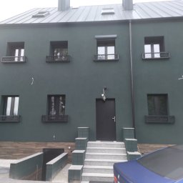 S/Mdd-Bud - Nowoczesny dom z ciemnozieloną elewacją, metalową blachą na dachu, czarnymi oknami z ozdobnymi balustradami i schodami wejściowymi wyłożonymi jasną kostką, częściowo zasłonięty niebieskim samochodem.