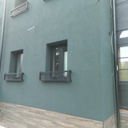 S/Mdd-Bud - Elewacja budynku z zielonym tynkiem strukturalnym, dekoracyjnymi balustradami okiennymi i okładziną imitującą drewno na dole ściany.