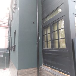 S/Mdd-Bud - Fragment elewacji budynku z ciemnozielonym tynkiem i drewnopodobną okładziną cokołu; widoczne okno z czarną ramą i pionowa rura spustowa.