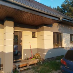 S/Mdd-Bud - Dwukolorowa elewacja domu jednorodzinnego z widoczną częścią dachu, drewnianym podbitką i schodami wejściowymi; obok wejścia stoi czerwona zmiotka i buty robocze.