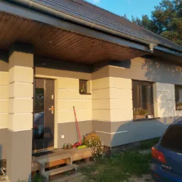 Dwukolorowa elewacja domu jednorodzinnego z widoczną częścią dachu, drewnianym podbitką i schodami wejściowymi; obok wejścia stoi czerwona zmiotka i buty robocze.