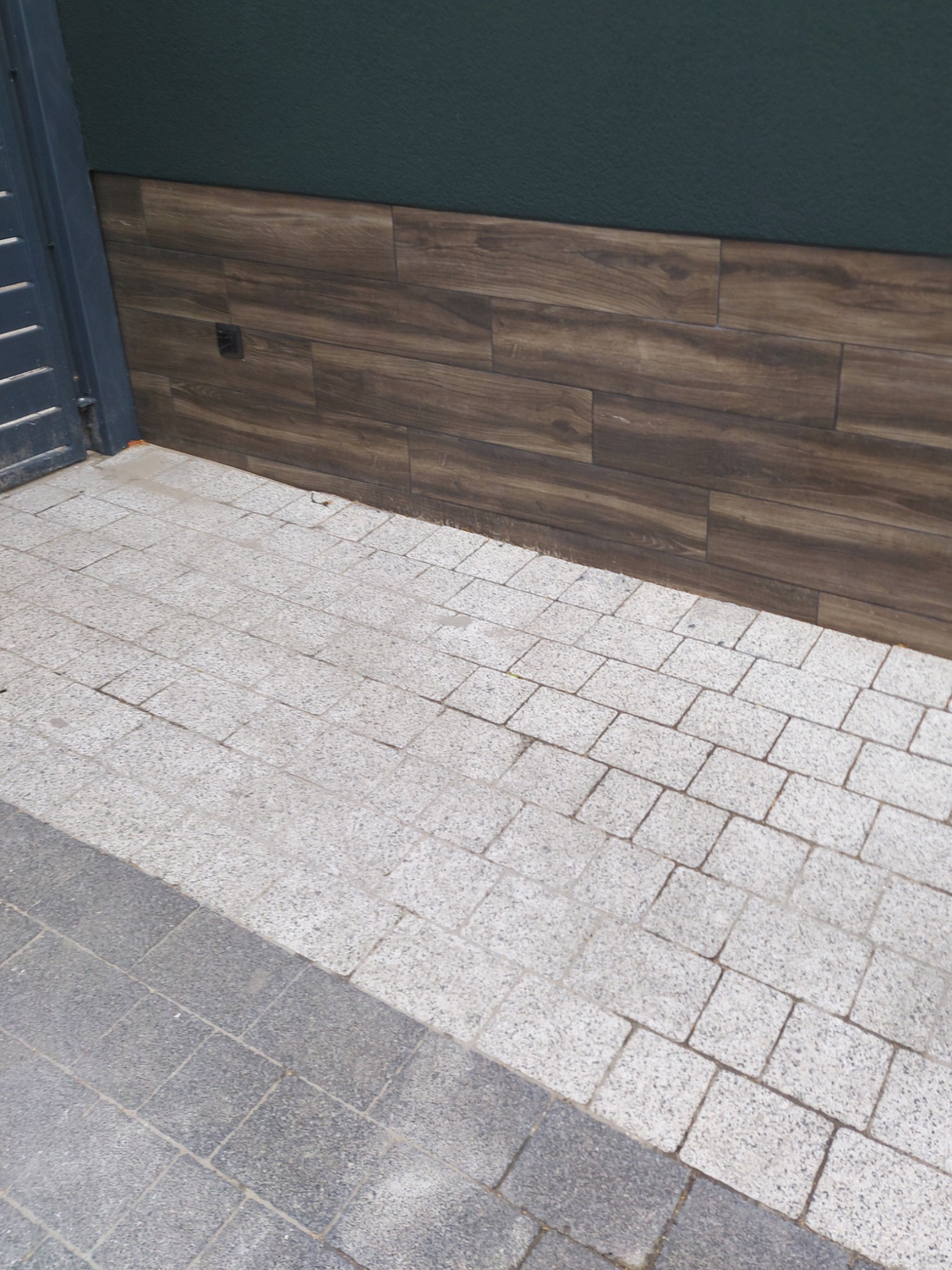 Fragment elewacji budynku z ciemnozielonym tynkiem i panelami imitującymi drewno, przylegający do chodnika wyłożonego szarą kostką brukową.