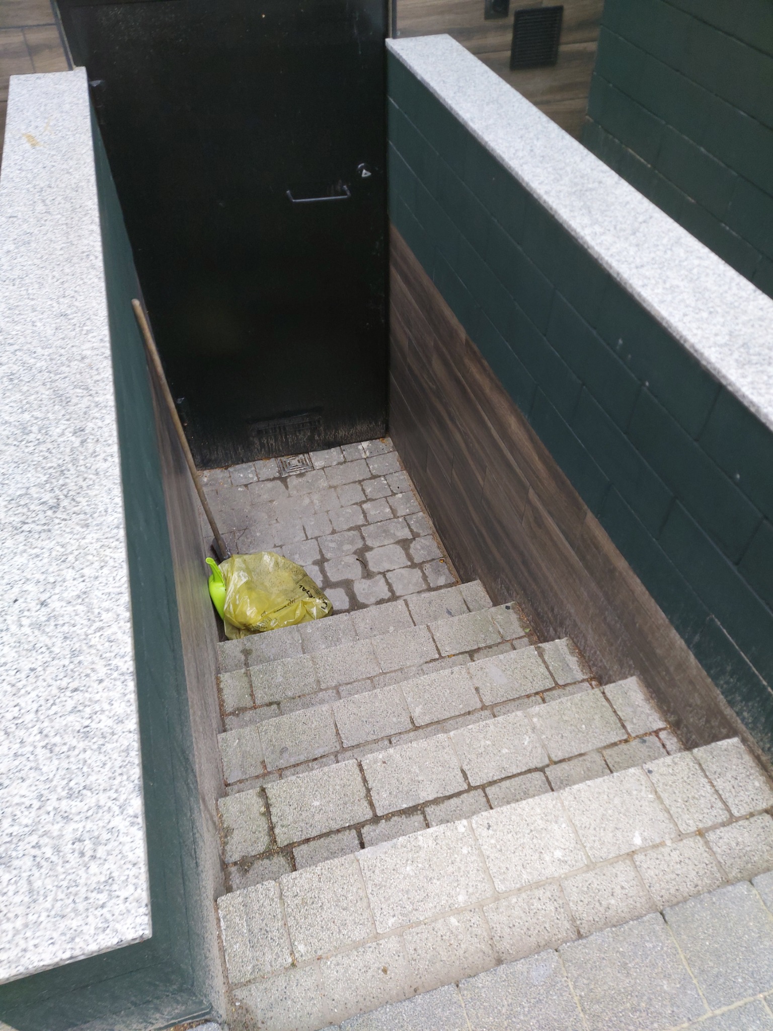 Zewnętrzne schody z szarej kostki prowadzące do czarnych drzwi piwnicznych, obok ściana wyłożona panelami imitującymi drewno, przy schodach leży żółta torba z odpadami.