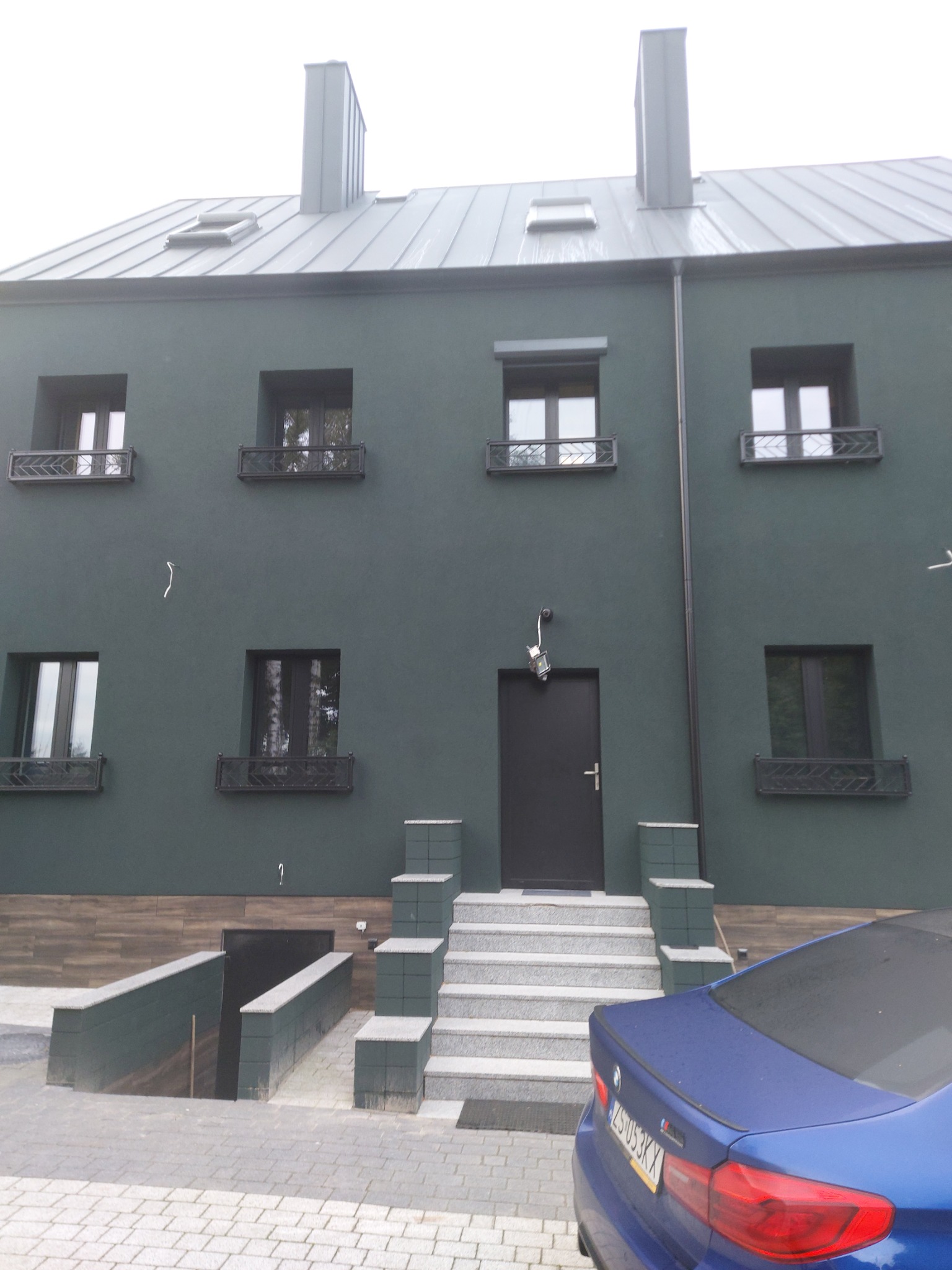 Nowoczesny dom z ciemnozieloną elewacją, metalową blachą na dachu, czarnymi oknami z ozdobnymi balustradami i schodami wejściowymi wyłożonymi jasną kostką, częściowo zasłonięty niebieskim samochodem.