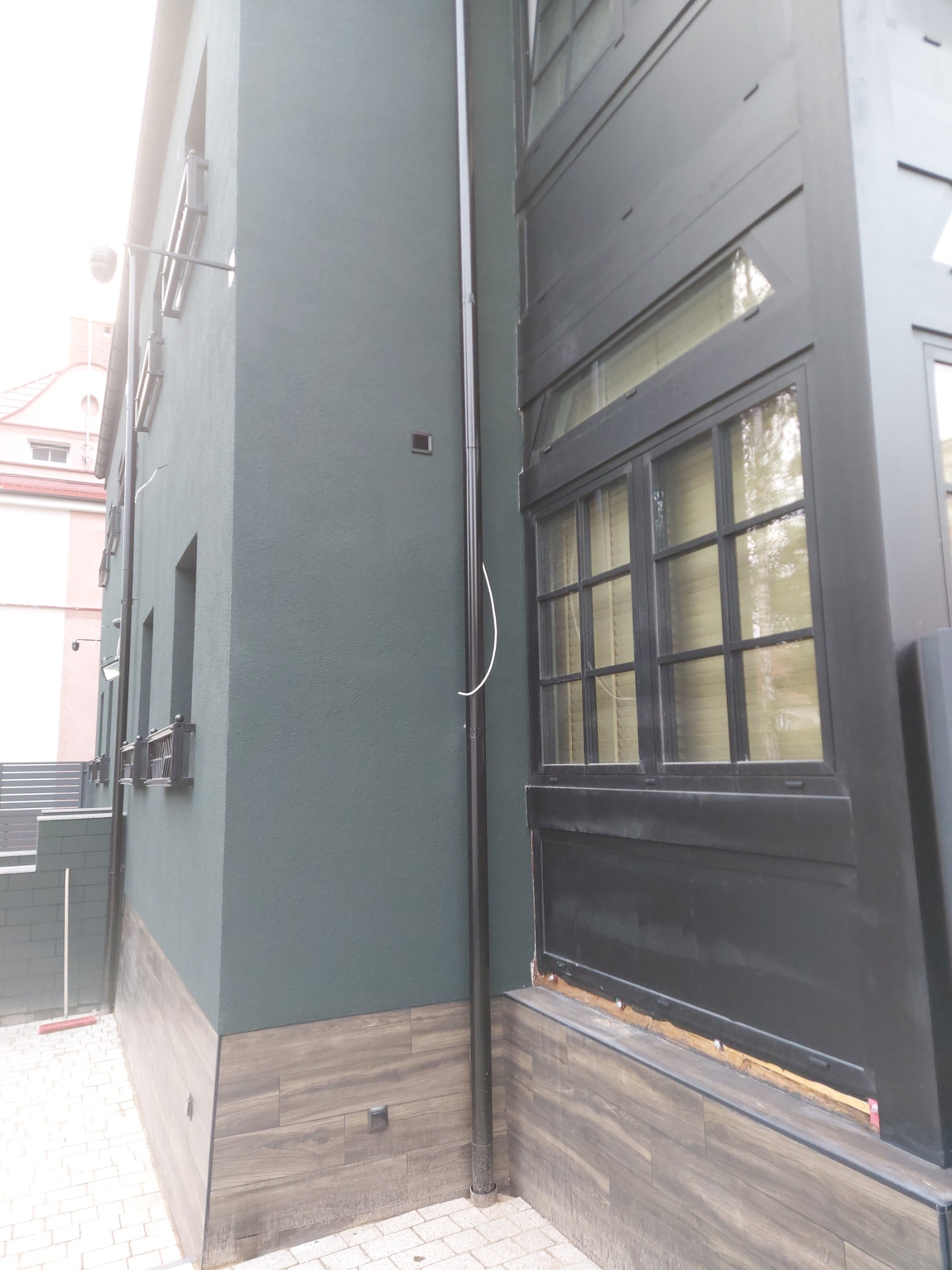 Fragment elewacji budynku z ciemnozielonym tynkiem i drewnopodobną okładziną cokołu; widoczne okno z czarną ramą i pionowa rura spustowa.