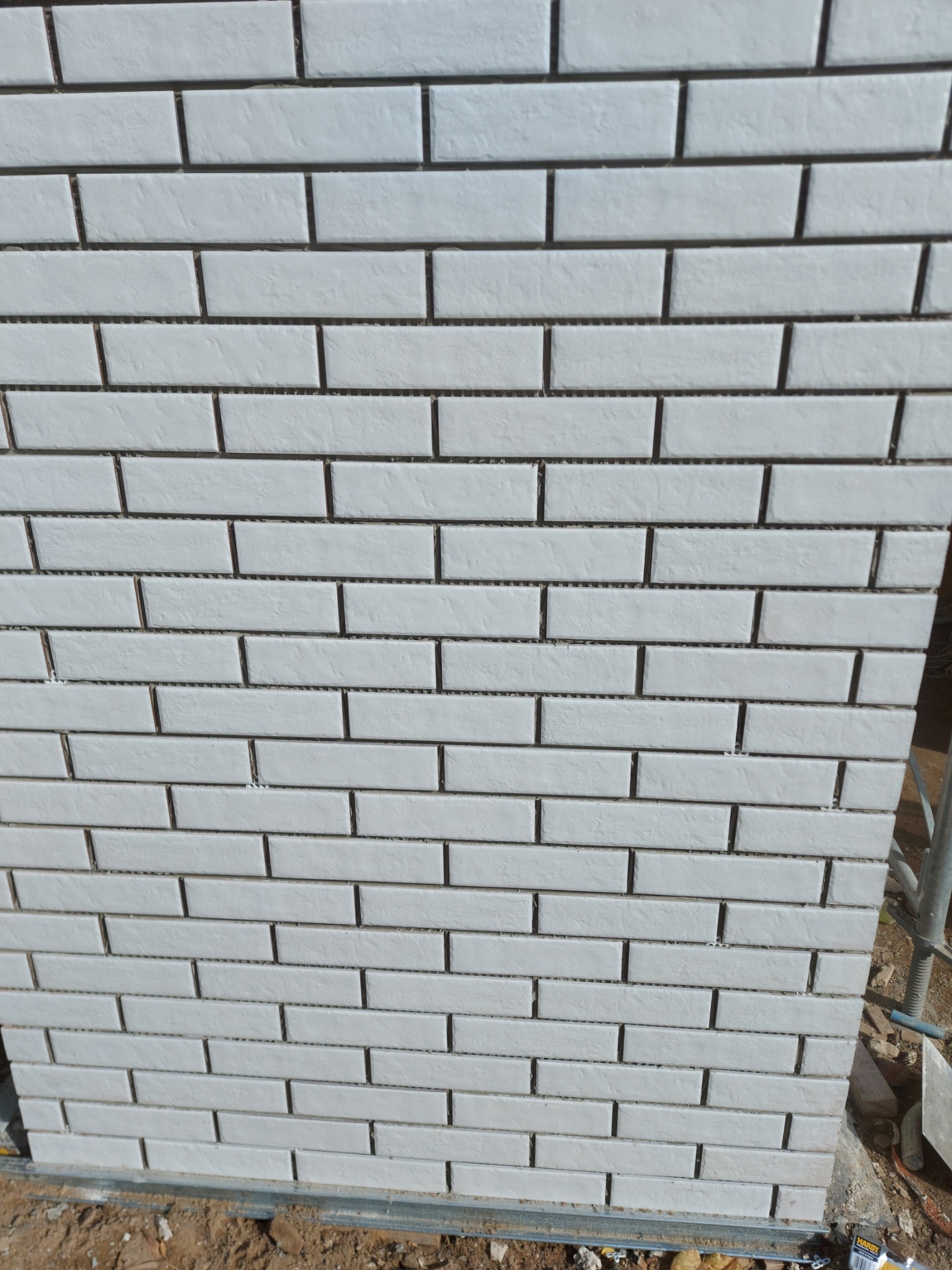 Fragment ściany pokrytej białymi, wąskimi płytkami klinkierowymi, ułożonymi w regularny wzór cegiełki, z widocznymi szarymi fugami i elementami rusztowania w tle.