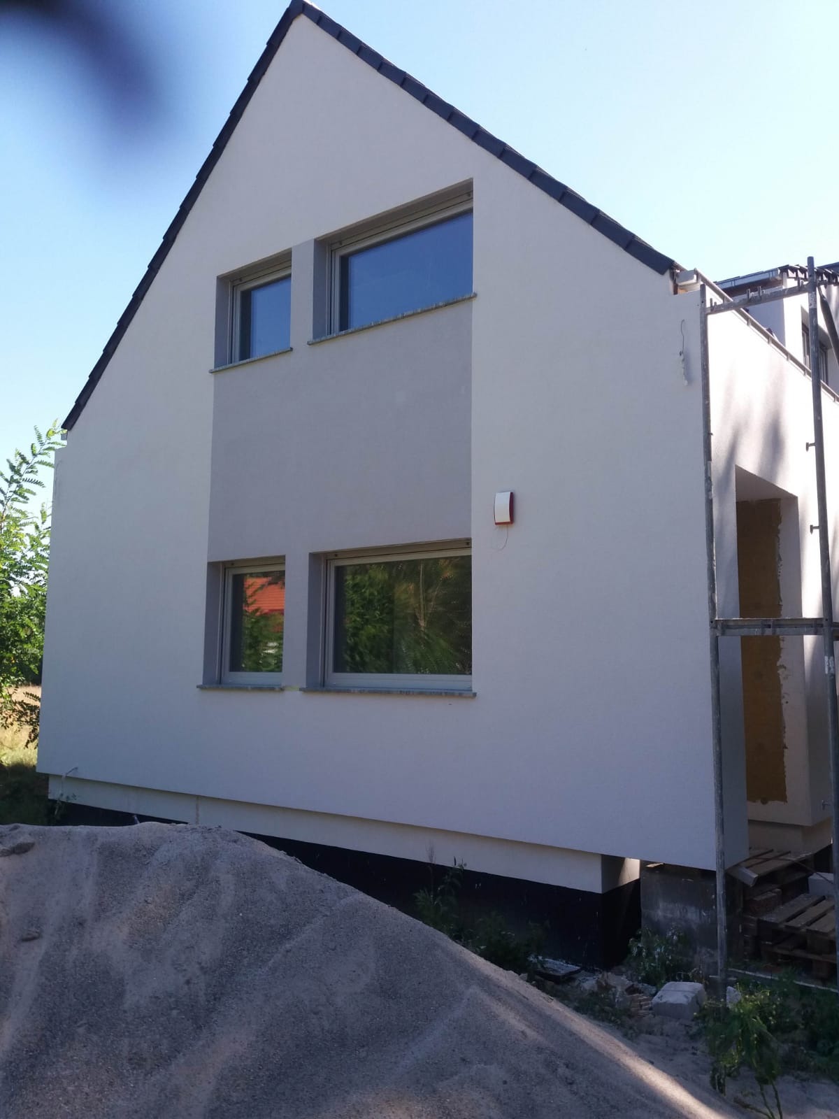 Jasnoszary dom jednorodzinny z ciemną dachówką w trakcie prac wykończeniowych elewacji, z widocznym rusztowaniem i stertą piasku u podstawy.