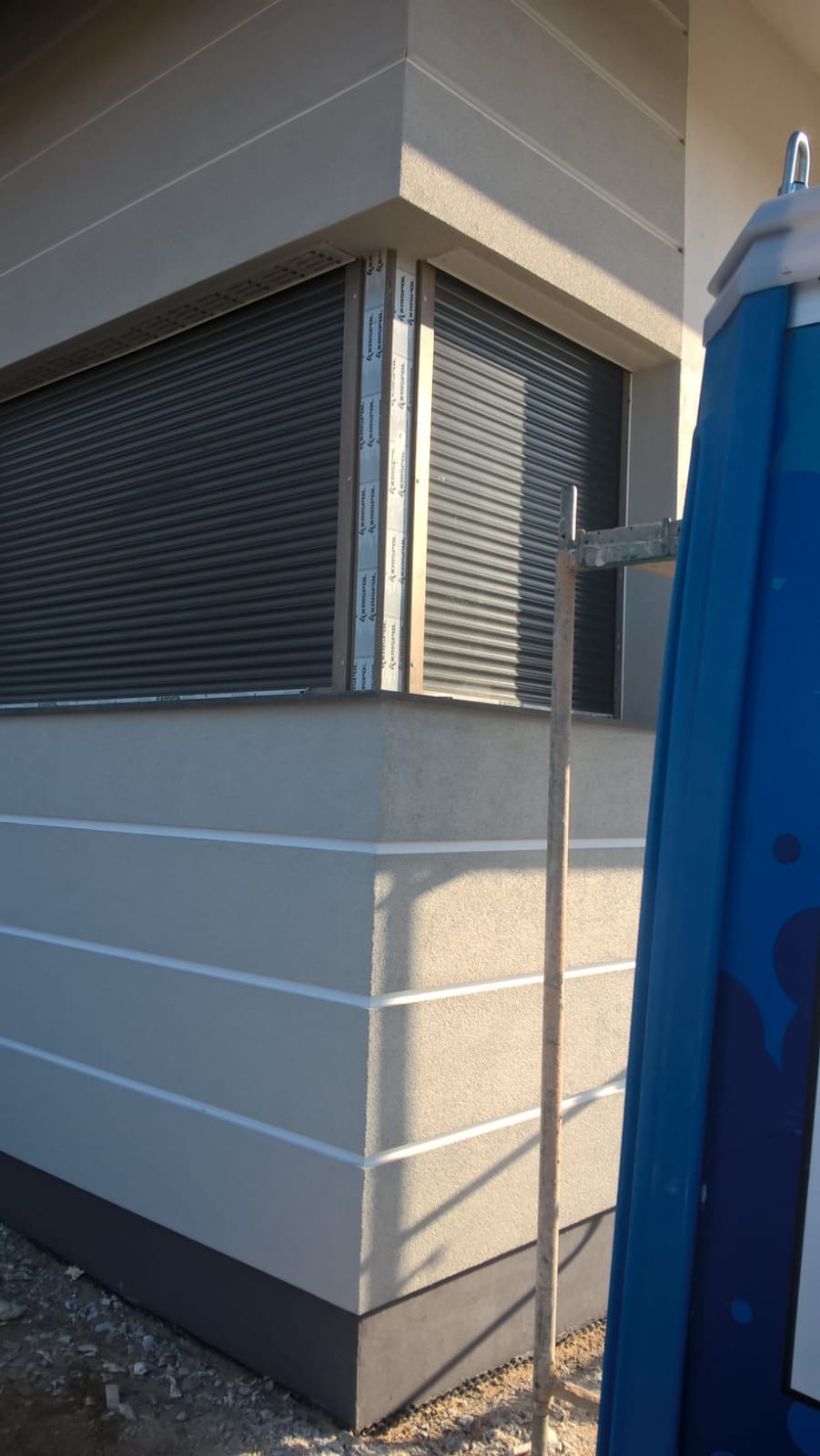 Narożnik budynku z szarą elewacją, ozdobiony białymi poziomymi paskami, z oknami z roletami zewnętrznymi. Na pierwszym planie widoczna niebieska toaleta przenośna i fragment rusztowania.