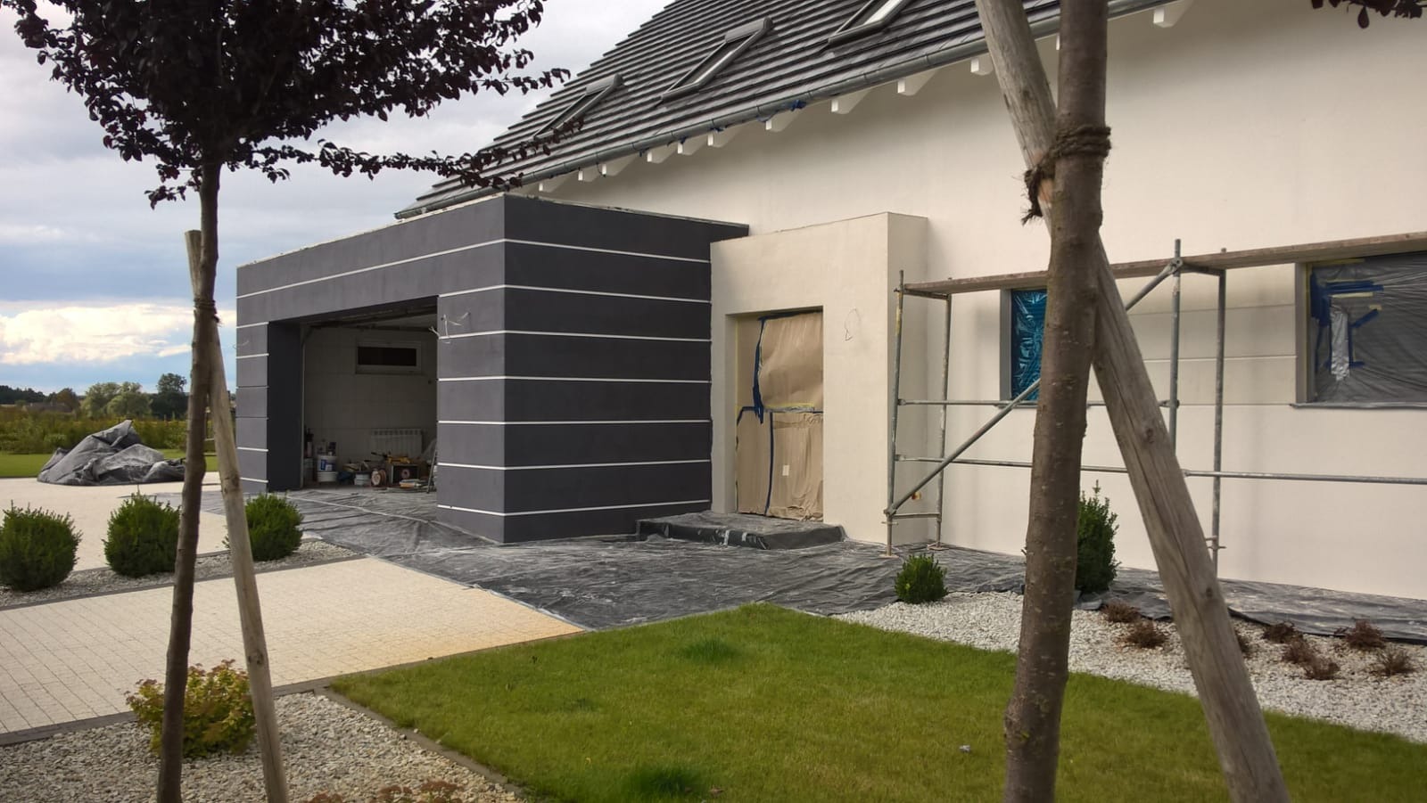 Elewacja domu jednorodzinnego w trakcie prac wykończeniowych: część pomalowana na biało, część w ciemnym szarym z poziomymi białymi pasami, okna zabezpieczone folią, widoczne rusztowanie, teren...